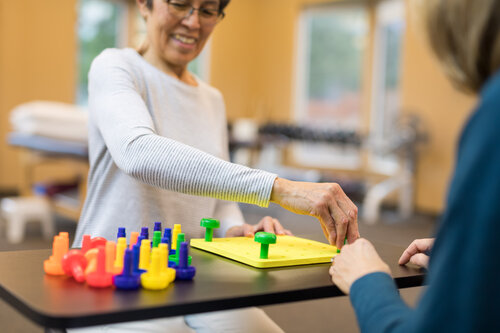 An elderly woman engages in a colorful pegboard activity, assisted by a caregiver. The scene is warm and supportive, set in a therapy room.