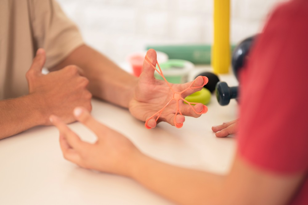 A therapist helps a child stretch an elastic band between their fingers at a table, practicing hand strength and fine motor skills in a calm session.