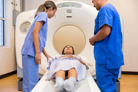 A patient in a hospital gown lies on a CT scan table, attended by two healthcare professionals in blue scrubs, inside a medical imaging room.