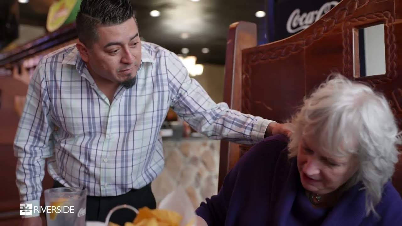 A waiter in a plaid shirt smiles warmly at an older woman seated in a restaurant booth. The atmosphere is cozy and inviting, suggesting friendly interaction.