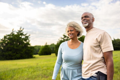 A smiling couple walks arm in arm through a lush green field under a partly cloudy sky, conveying a sense of joy and serenity in a natural setting.