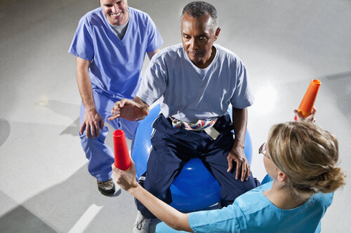 A man sits on a blue exercise ball, focusing on orange cones held by two healthcare professionals in scrubs, conveying a supportive rehabilitation setting.
