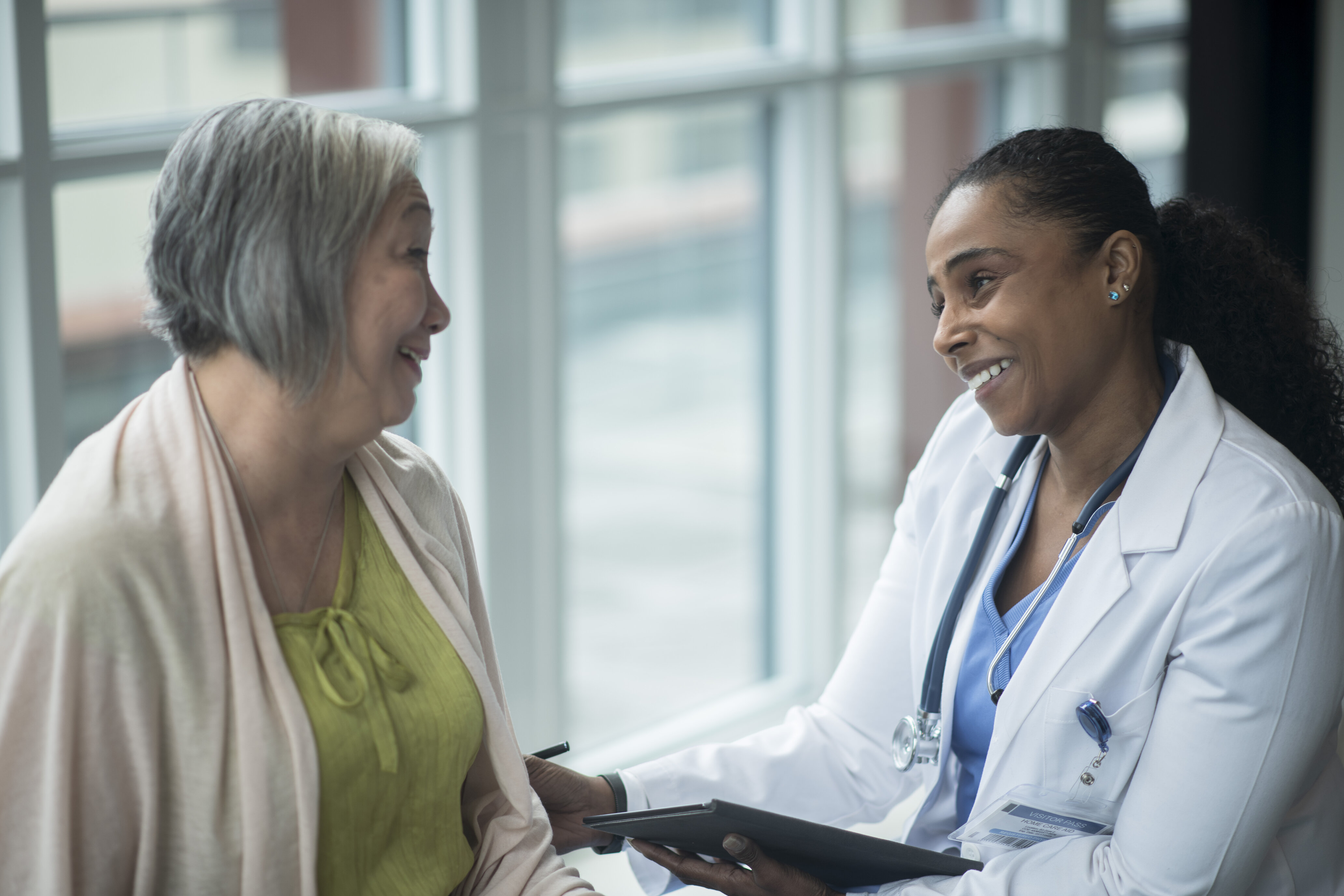 A doctor in a white coat and stethoscope warmly smiles and talks with a senior woman by a large window, creating an empathetic and caring atmosphere.