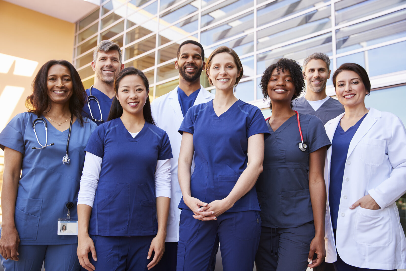 A diverse group of smiling healthcare professionals in blue scrubs and white coats stand together outdoors beside a modern hospital building, exuding teamwork.