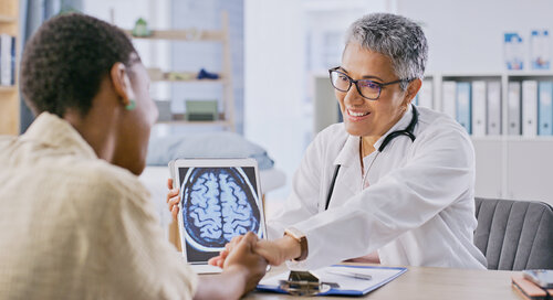 A doctor with short gray hair and glasses shows a brain scan to a patient in an office. The doctor is smiling, conveying a reassuring tone.