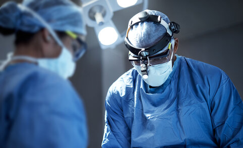 Surgeons in blue scrubs and masks operate under bright surgical lights. The focus is on a surgeon wearing magnifying eyewear, conveying concentration and precision.