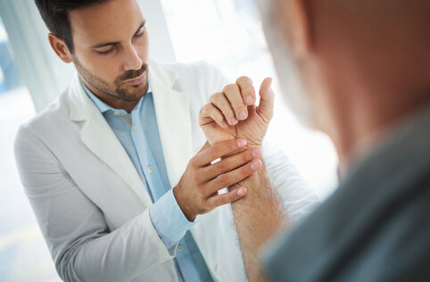 A doctor in a white coat gently examines a patient's wrist in a bright medical office. The scene conveys a sense of care and attentiveness.