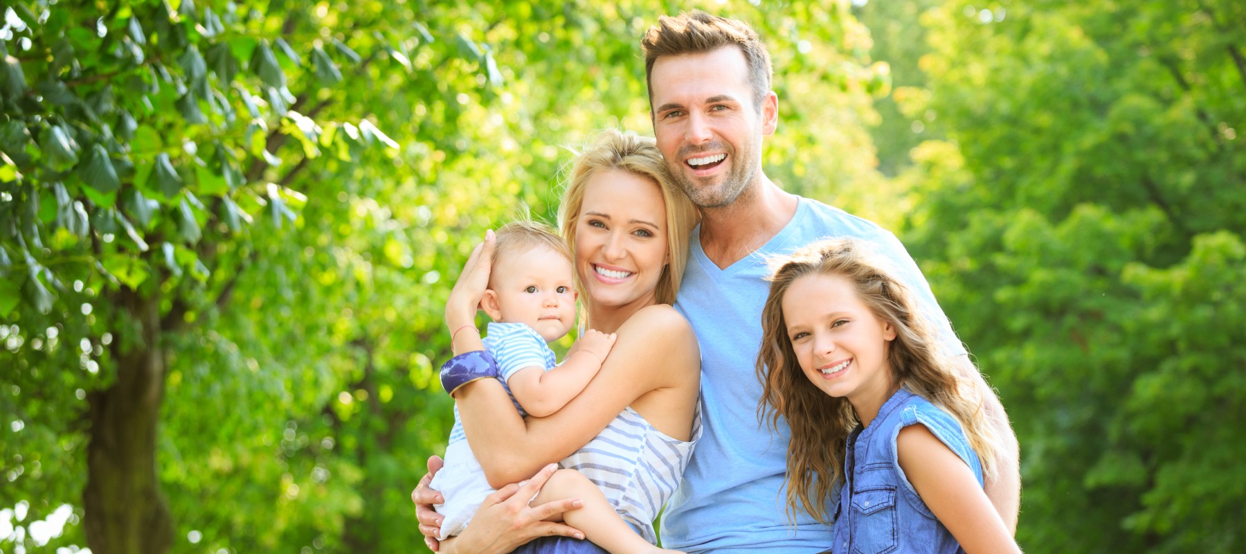 Smiling family of four, with parents holding a baby and a young girl standing beside them in a sunlit park. Green trees create a lively backdrop.