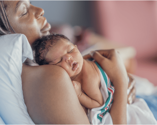 A new Mother, of African decent, holds her baby on her shoulder as she sits up in her hospital bed and they spend time bonding skin-to-skin.