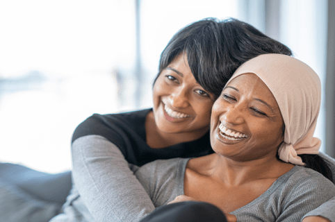 A young girl embraces her Mother who is fighting Cancer, as they share a close moment at home together. They are seated on a sofa and embracing one another. Both are dressed casually and smiling as they enjoy a laugh.