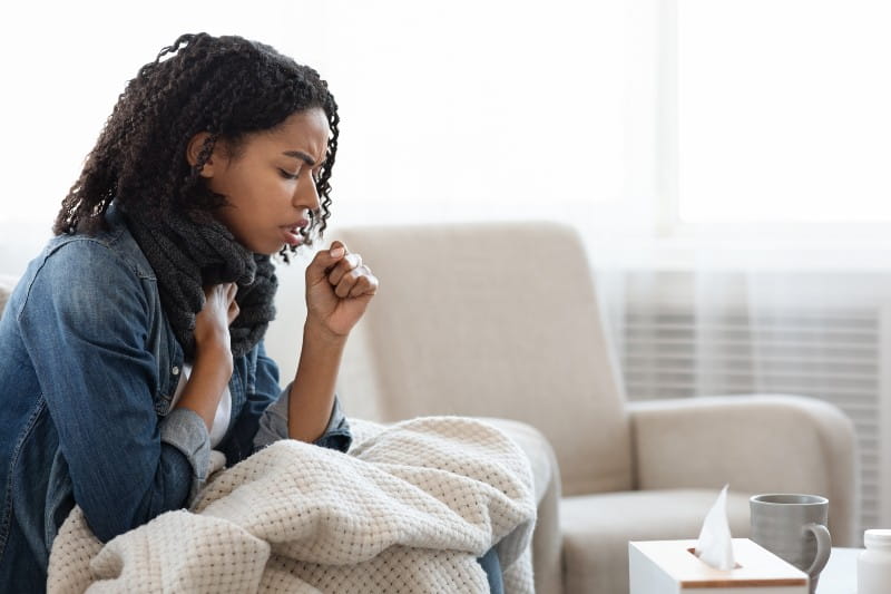 Black Woman Coughing Hard At Home, Sitting On Couch Wearing Scarf And Covered With Blanket