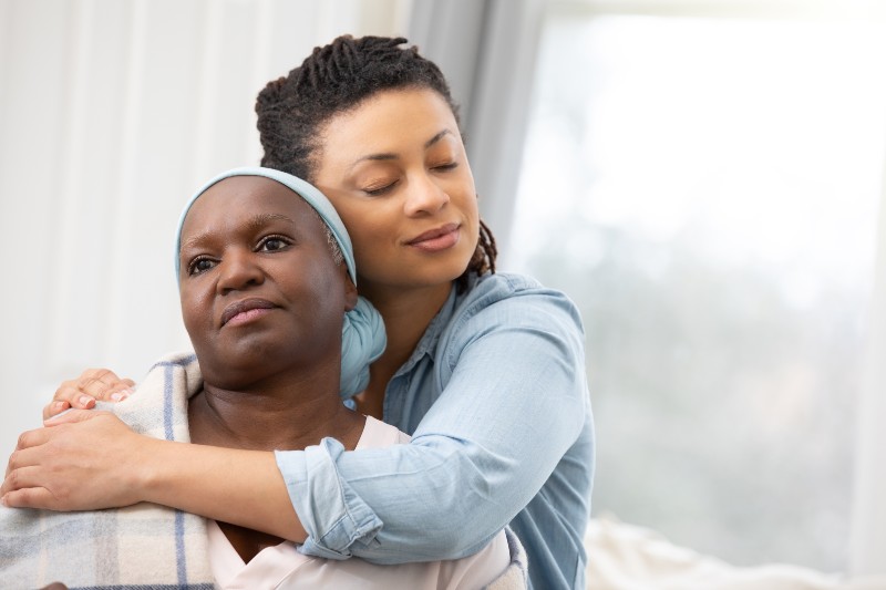 A senior woman looks away, while daughter closes her eyes and gives a caring hug