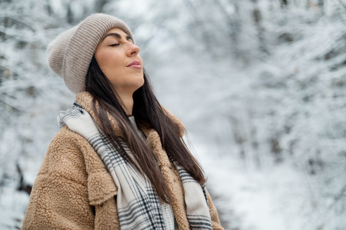 Woman in warm winter clothing stands outdoors in a snowy forest, eyes closed, expressing serene contentment. Trees and path covered in snow.