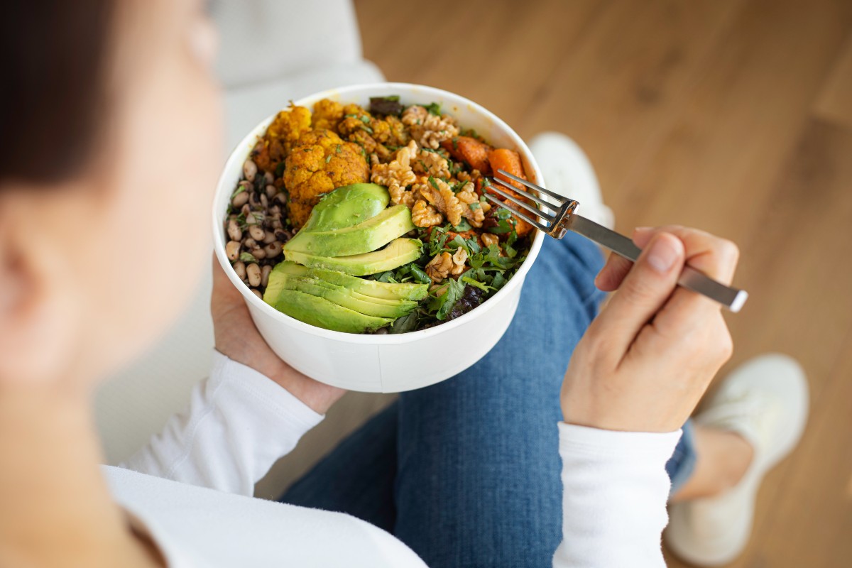 A person holds a bowl of colorful salad featuring sliced avocado, cauliflower, greens, and walnuts, conveying a sense of healthy eating.