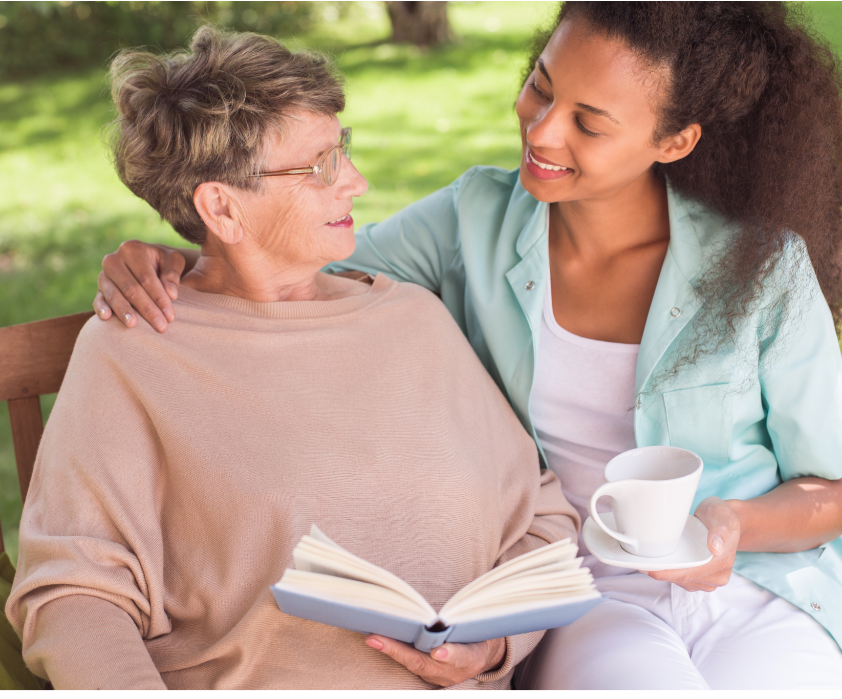 Two women sit on a bench in a garden. The older woman holds a book, while the younger woman, smiling, holds a cup of tea, exuding warmth and companionship.
