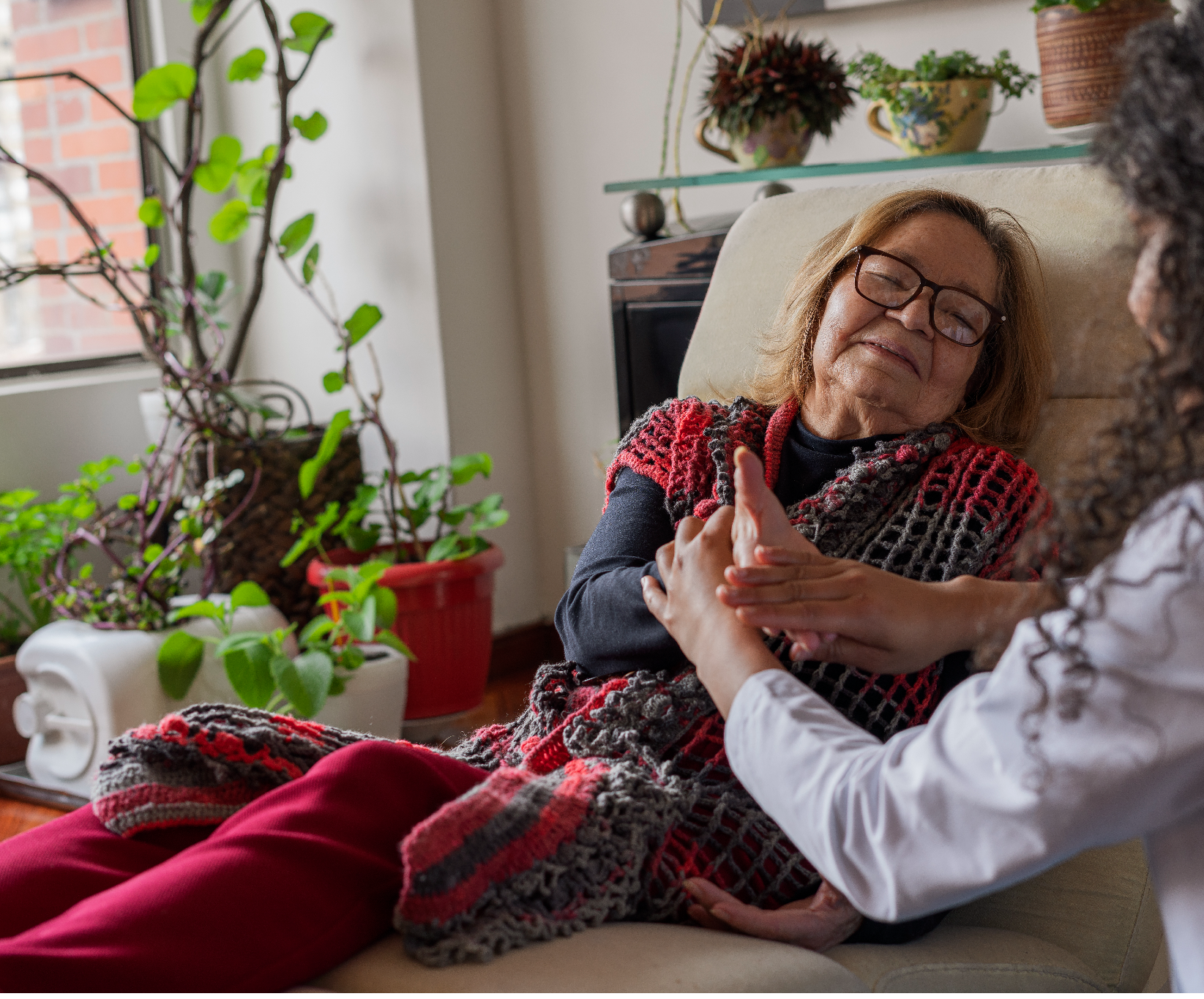 An elderly woman with glasses, wrapped in a red and gray blanket, smiles contentedly as a caregiver massages her hand in a cozy, plant-filled room.