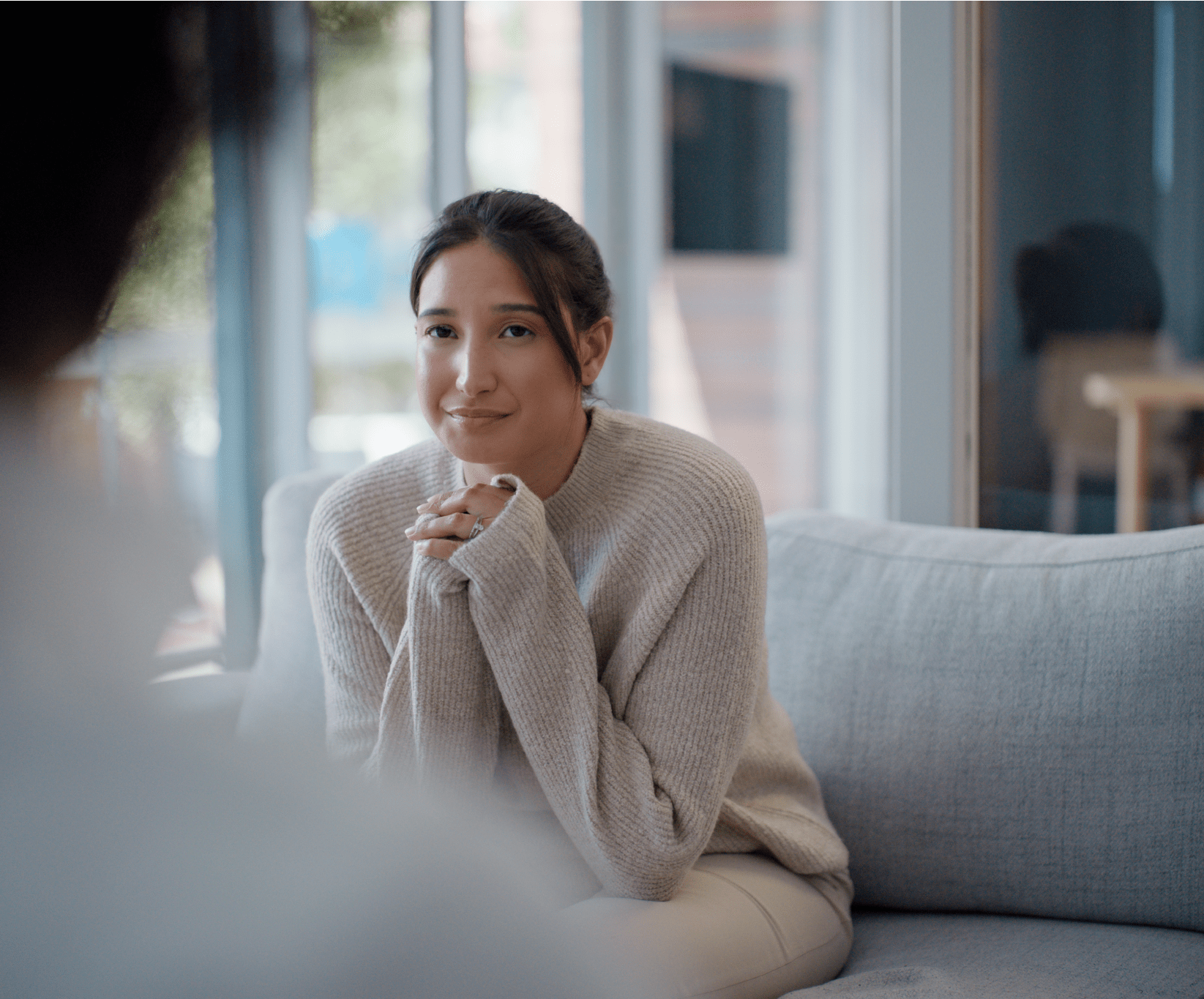 A woman in a cozy sweater sits on a gray sofa, smiling softly, with sunlight streaming through large windows behind her, creating a calm atmosphere.