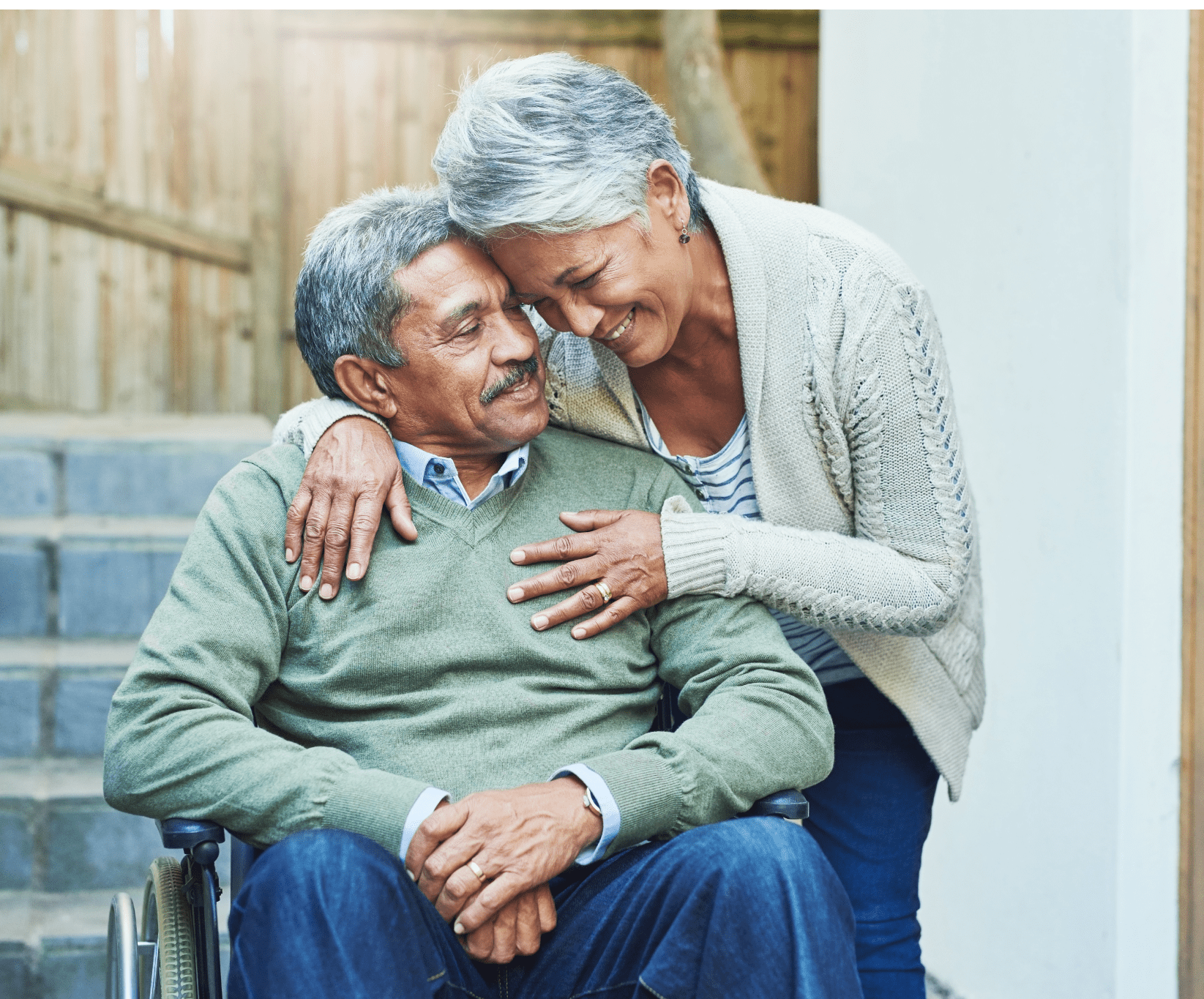 An elderly couple shares a tender moment outdoors. The man sits in a wheelchair, smiling, as the woman leans over, embracing him warmly. Emotional and loving scene.