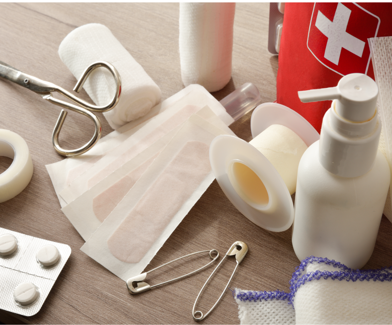 First aid supplies on a wooden table, including bandages, scissors, adhesive tape, safety pins, a spray bottle, and a red medical kit.