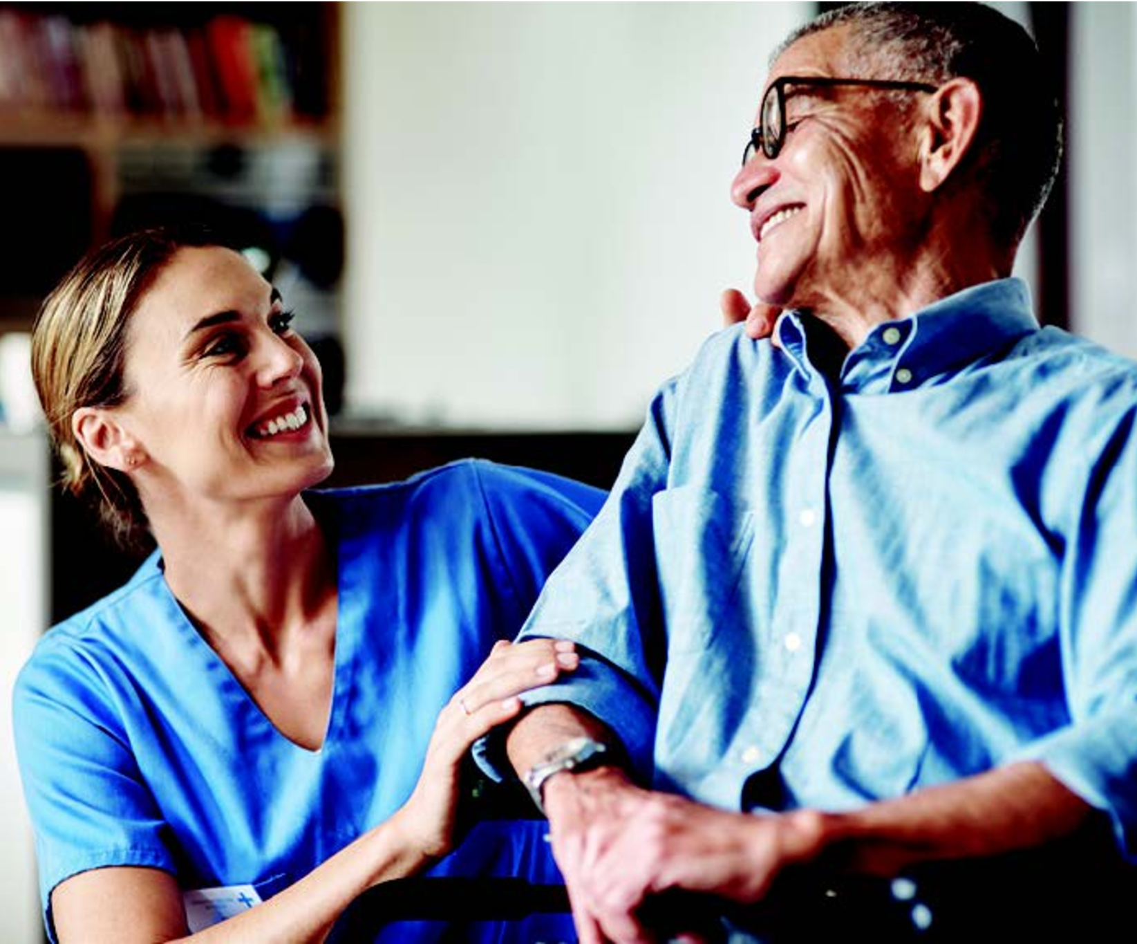 A caregiver in blue scrubs and an elderly man in glasses share a warm smile. The setting is cozy and conveys a sense of trust and companionship.