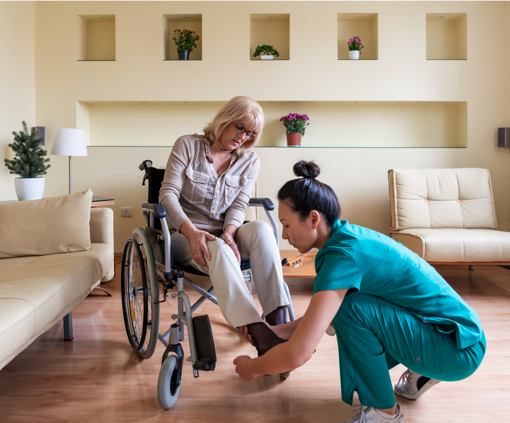 A caregiver in teal scrubs helps a woman in a wheelchair with her shoe in a cozy living room. The mood is caring and supportive, with plants and soft lighting.