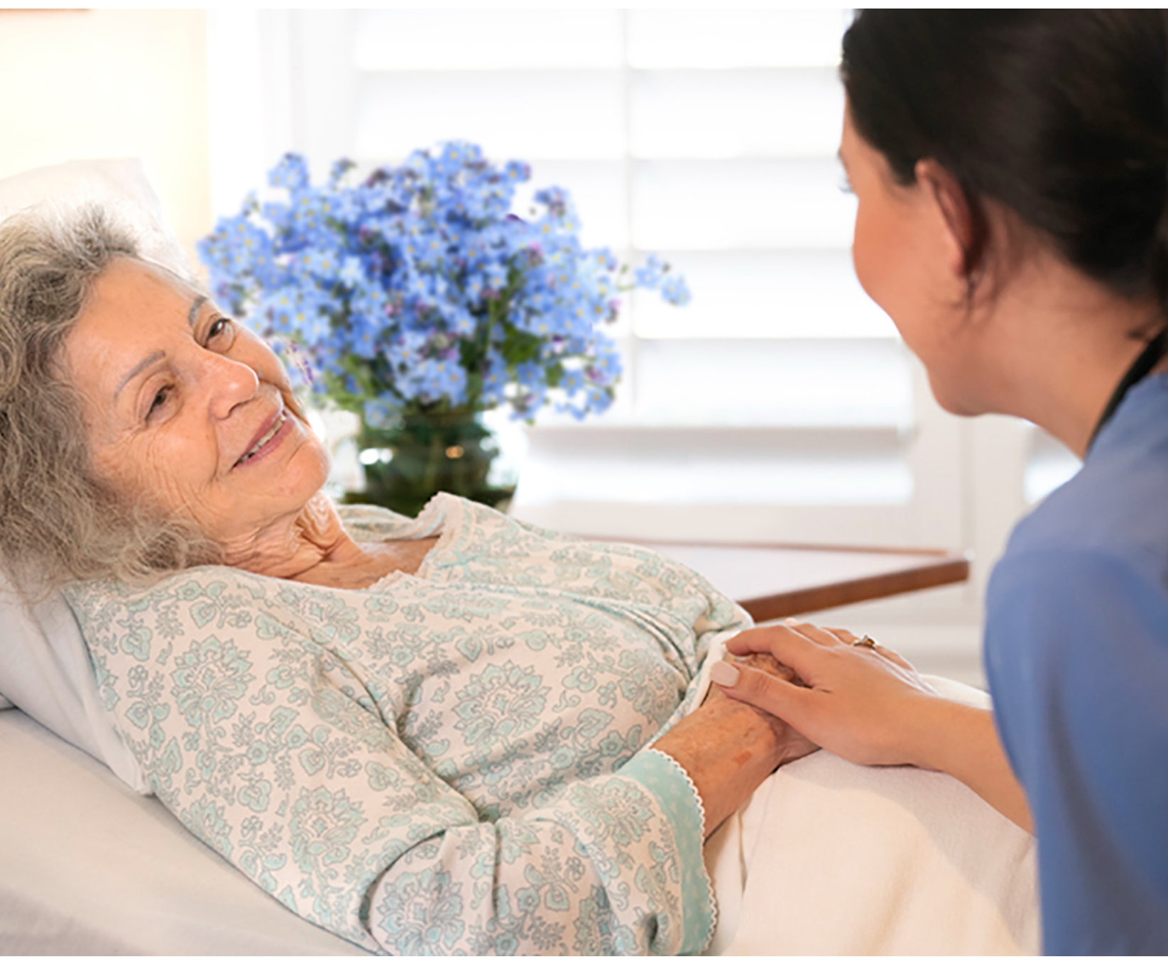 Elderly woman in bed smiles at a caregiver holding her hand. Blue flowers in the background. Scene conveys warmth and compassion.