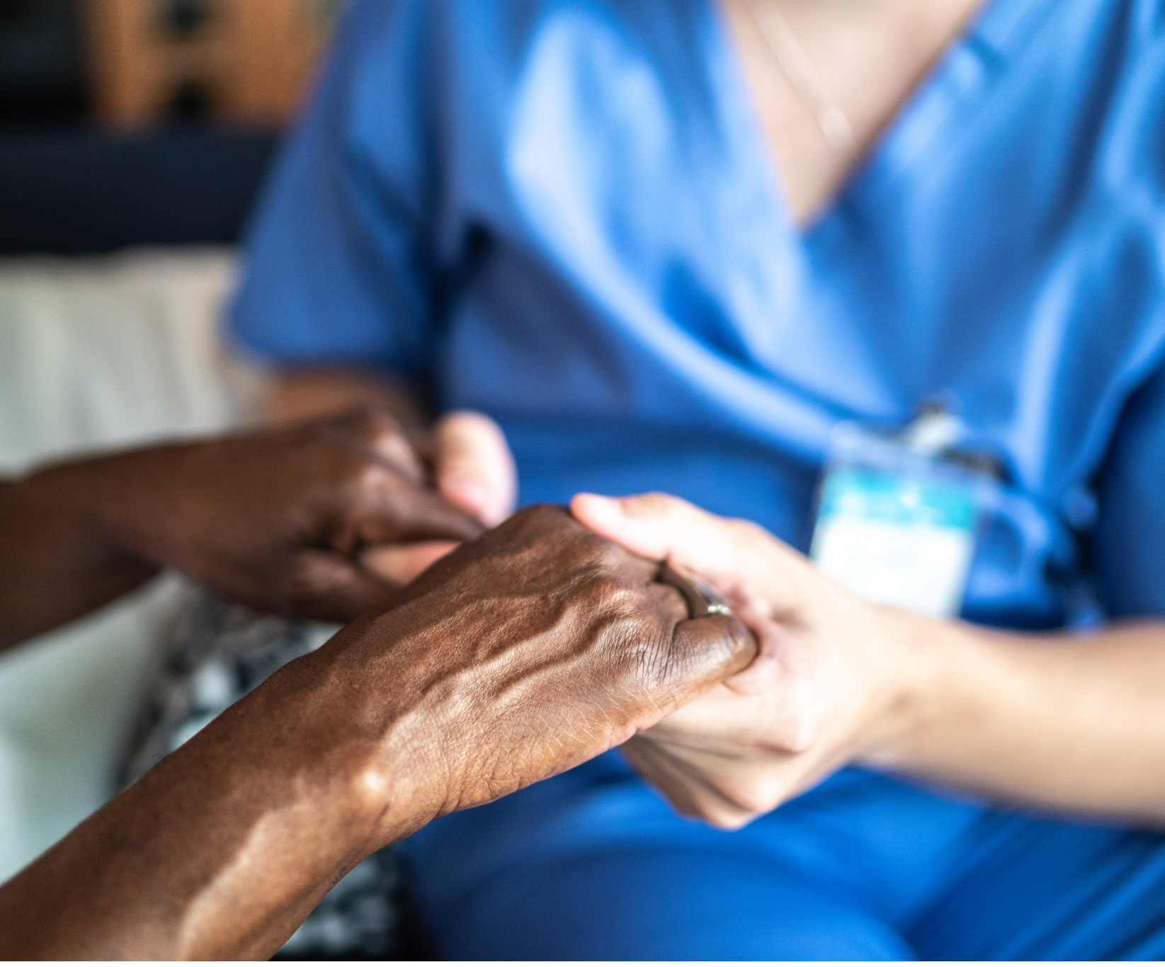 A nurse in blue scrubs gently holds the hand of an elderly person, conveying care and compassion in a healthcare or supportive setting.