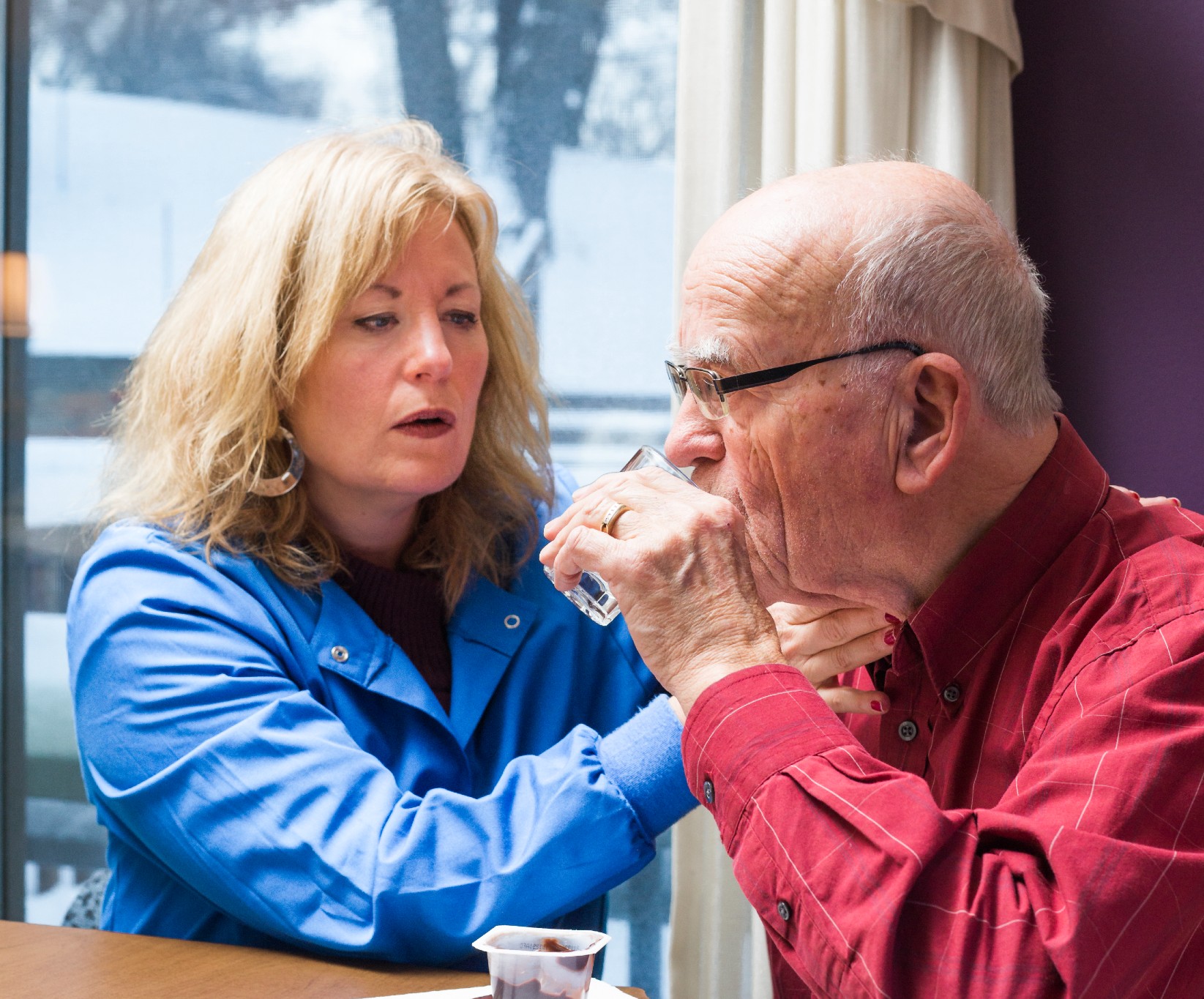 A woman in a blue coat supports an elderly man in glasses as he drinks from a glass indoors. The setting suggests care and support, with a snowy view outside.