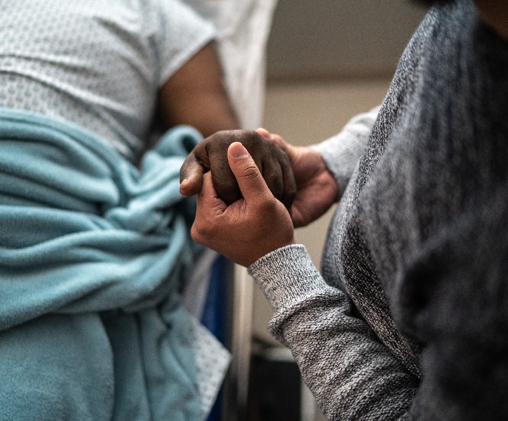 A person in a gray sweater gently holds the hand of someone in a hospital bed, covered with a light blue blanket, conveying a sense of support and care.