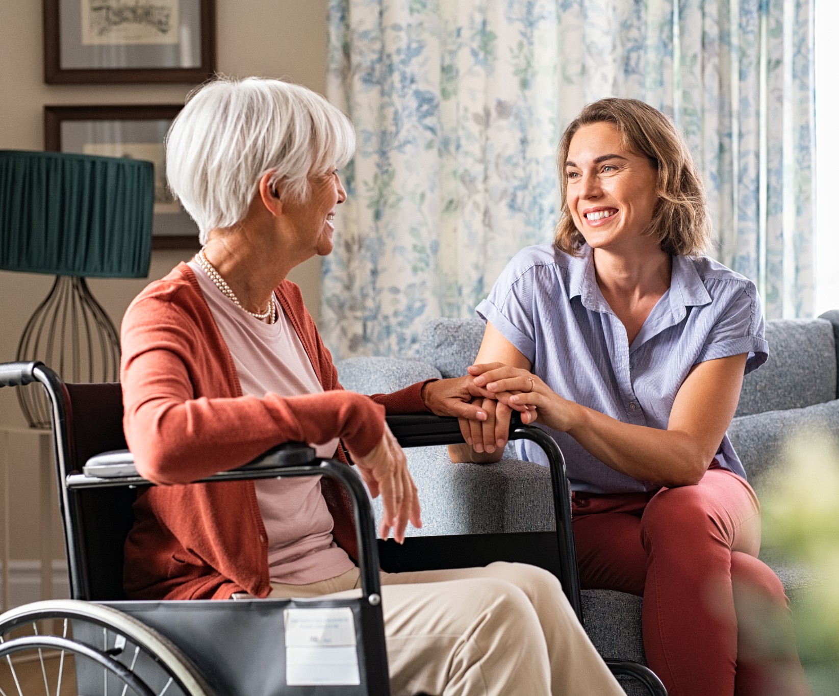 A senior woman in a wheelchair smiles at a younger woman sitting beside her on a couch. They hold hands warmly, sharing a joyful and supportive moment.
