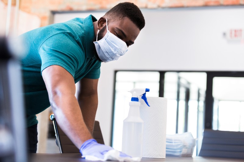 serious mid adult man cleans a conference table in his office while wearing a protective face mask