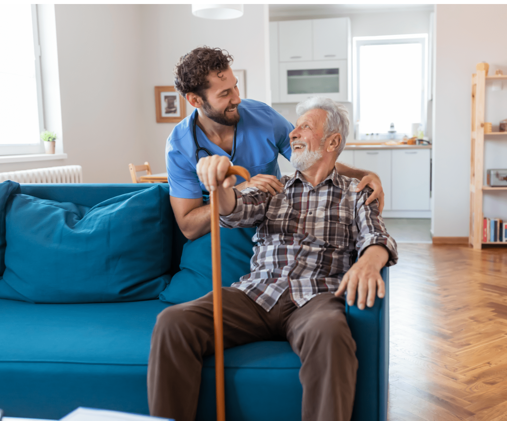 A caregiver in blue scrubs smiles warmly at a seated elderly man with a cane. They share a joyful moment in a cozy, sunlit living room.