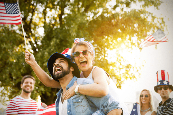 Young adults celebrate outdoors with American flags. A woman joyfully piggybacks a man, both smiling. The scene is sunny and festive, evoking patriotism.