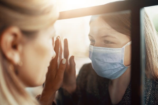 Mother and daughter are separated by glass, one wearing a mask