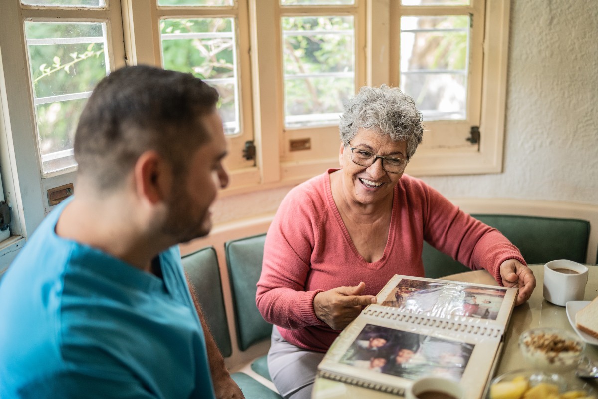 A smiling elderly woman in a pink sweater shares a photo album with a man in a blue shirt at a cozy table by a window. The mood is warm and nostalgic.