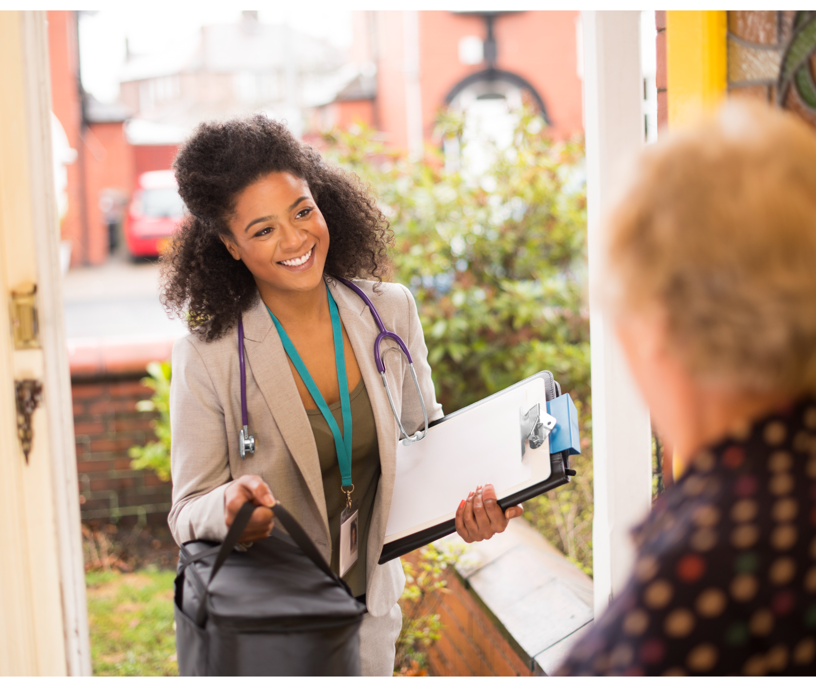 Healthcare worker with stethoscope carrying medical bag and clipboard, visiting a patient at home and speaking at the front door.