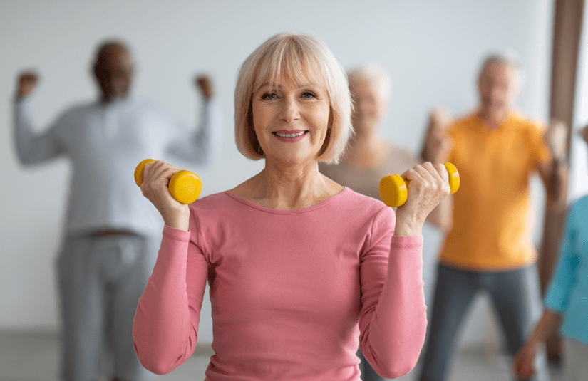 Elderly woman in a pink top lifts yellow dumbbells, smiling confidently, while a diverse group of seniors exercises in the background. Bright and lively atmosphere.